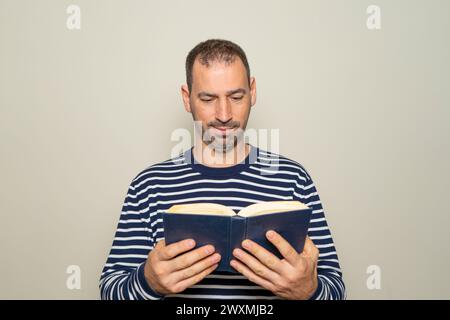 Homme hispanique avec une barbe dans ses années 40 portant un pull rayé lisant un livre en papier, il est concentré et excité par le magnifique contenu de la Banque D'Images