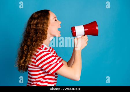 Photo de profil latéral d'une femme adorable avec une coiffure ondulée habillée t-shirt rayé criant dans un haut-parleur isolé sur fond de couleur bleue Banque D'Images