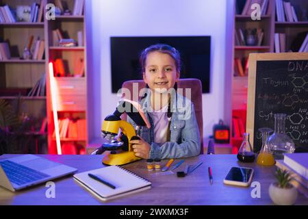 Portrait de petite fille souriante assise à table et regarde dans un microscope électronique à l'aide d'un smartphone. Enfant caucasien dans les études de vêtements décontractés par d Banque D'Images