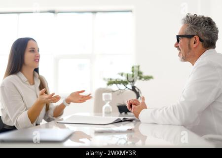 Un médecin senior aux cheveux gris, portant des lunettes et une blouse blanche, écoute attentivement une jeune patiente. Caractère communicatif et professionnel de la consultation médicale Banque D'Images