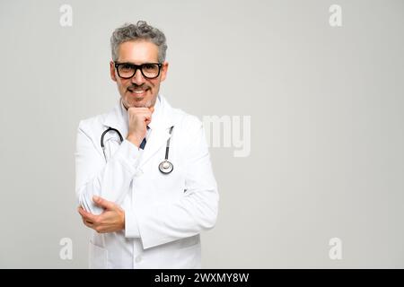 Un médecin chevronné aux cheveux gris se tient confiant avec un stéthoscope autour du cou, souriant chaleureusement à la caméra, dégageant un sentiment de confiance et de professionnalisme. Banque D'Images