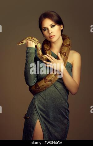 Jeune femme en robe verte pailletée avec maquillage, posant avec un serpent sur fond brun de studio. Banque D'Images