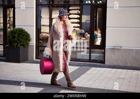 Femme mature à la mode marchant dans la ville, vêtue d'un long manteau beige, de bottes cosaques, d'un chapeau avec imprimé animal et d'une boîte de sac à main originale. style de rue Banque D'Images