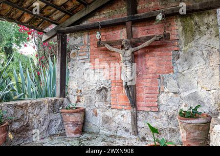 Énorme vieille croix en bois avec Christ crucifié dans le hangar de ranch, toit de tuiles, murs de briques usés, pots en argile avec de petites plantes vertes, bougainvilliers rouges et tr Banque D'Images