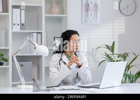 Médecin féminin hispanique réfléchi assis à son bureau dans un bureau médical lumineux, regardant un écran d'ordinateur portable. Banque D'Images