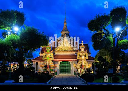 Entrée principale du temple Wat Arun avec deux statues de démons sur le devant. Le bâtiment est éclairé la nuit. Bangkok Banque D'Images