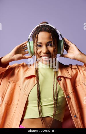 heureuse fille afro-américaine dans les années 20 portant des écouteurs et une tenue décontractée vibrante sur fond violet Banque D'Images