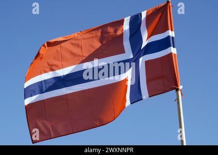 Un drapeau rouge vif avec une croix bleue encadrée en blanc, représentant la Norvège, flotte dans la brise sous un ciel sans nuages. Banque D'Images