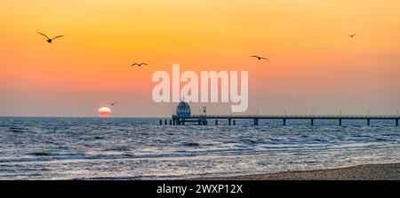 Lever de soleil orange sur la jetée de la mer Baltique à Zinnowitz sur Usedom, avec des mouettes volant au-dessus de la mer. Banque D'Images