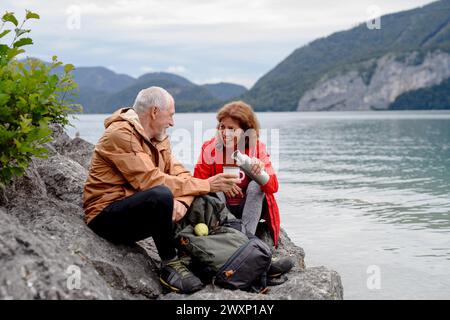 Couple de personnes âgées actives randonnant ensemble dans les montagnes. Boire du café et avoir une collation saine, pour obtenir de l'énergie. Touriste senior avec des sacs à dos au repos Banque D'Images