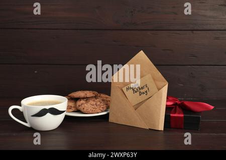 Carte avec phrase bonne fête des pères dans l'enveloppe, biscuits, tasse avec moustache et boîte cadeau sur table en bois Banque D'Images