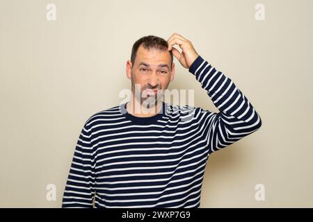 Homme hispanique barbu dans ses années 40 portant un pull rayé se grattant la tête tout en essayant de prendre une décision, doute concept. Isolé sur studio beige Banque D'Images