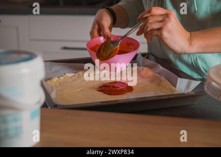 La femme verse de la sauce tomate faite maison sur la pâte à pizza tartinée dans une poêle. Pizza maison se préparant à être cuite à la maison Banque D'Images