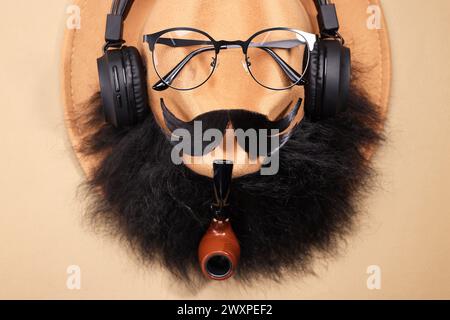 Visage d'homme en moustache artificielle, barbe, lunettes et chapeau sur fond beige, vue de dessus Banque D'Images