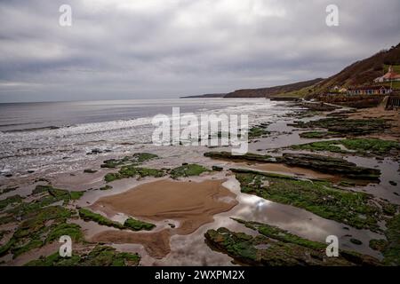 Une plage de sable avec des rochers couverts d'algues et un ciel gris sombre dominé par des cabanes de plage, Scarborough South Bay, North Yorkshire, Angleterre. Banque D'Images