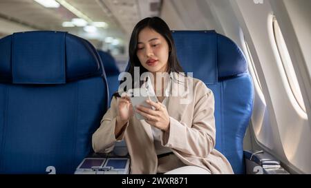 Une femme d'affaires asiatique attrayante dans un costume d'affaires vérifie les messages ou allume le mode avion sur son smartphone pendant le vol ou avant t Banque D'Images