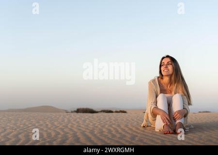 Plein corps vue latérale de jeune touriste souriante pieds nus dans des vêtements décontractés regardant loin tout en étant assis avec les jambes pliées sur une dune de sable dans la soirée da Banque D'Images