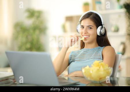 Femme avec ordinateur portable et casque mangeant des chips regardant la caméra à la maison Banque D'Images