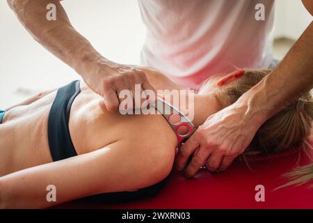 Thérapeute de massage de récolte faisant le massage avec l'outil IASTM sur l'épaule de la patiente couchée dans le centre de bien-être pendant le traitement de réadaptation à la thérapie Banque D'Images