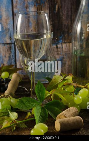 Verre de vin blanc sur un fond en bois avec des raisins, des feuilles et des bouchons de vin. Banque D'Images