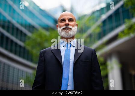 Homme d'affaires barbu mature dans un costume se tient en toute confiance à l'extérieur avec des immeubles de bureaux en arrière-plan Banque D'Images