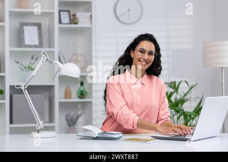 Femme hispanique professionnelle travaillant à la maison, regardant la caméra avec un sourire joyeux, bureau à domicile bien équipé en arrière-plan. Banque D'Images