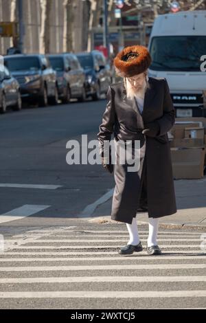 Un homme juif hassidique plus âgé portant de hauts bas blancs et un chapeau de fourrure shtreimel traverse la rue. Pendant Pourim 2023 à Brooklyn, New York. Banque D'Images