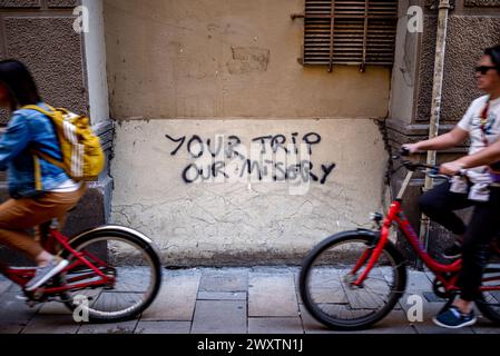 Dans le quartier gothique de Barcelone, un groupe de touristes en vélo passe un graffiti anti-tourisme lisant votre voyage notre misère. Des manifestations contre le tourisme de masse ont lieu depuis des années dans la capitale catalane. La Catalogne a eu un nombre record de visiteurs pour les vacances de Pâques 2024. Banque D'Images