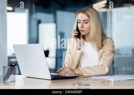 Une femme d'affaires blonde concentrée dans un blazer beige utilise un ordinateur portable et parle sur un smartphone dans un environnement de bureau moderne. Banque D'Images