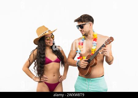 Charmant couple profitant d'une journée ensoleillée à la plage. femme latine dans un bikini sourit comme elle regarde son petit ami jouant du ukulélé, portant un ne de fleur Banque D'Images