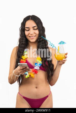 Une jeune femme latina portant un bikini est vue en train de texter sur son smartphone tout en tenant un soda orange d'été dans son autre main. Elle semble détendue et en Banque D'Images