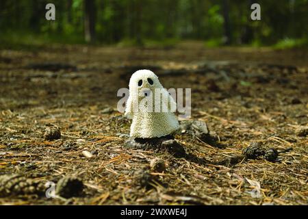 Un petit jouet fantôme blanc qui sourit et se tient sur le sol dans la forêt parmi les vieilles aiguilles et les cônes Banque D'Images