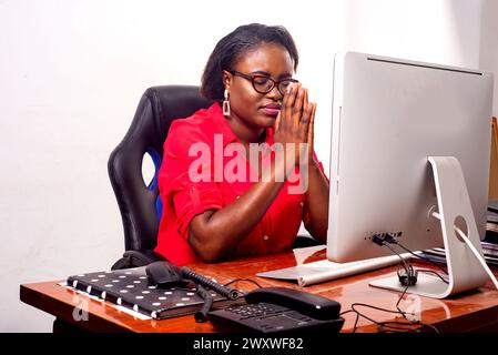 belle femme d'affaires adulte portant des lunettes optiques assis au bureau face à l'ordinateur portable et priant avec les mains ensemble. Banque D'Images