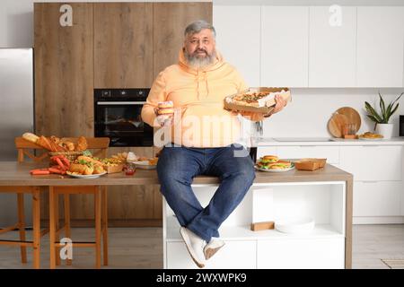 Homme mature en surpoids assis sur une table pleine de nourriture malsaine dans la cuisine. Concept de suralimentation Banque D'Images