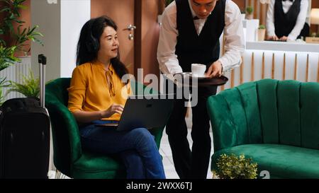 Femme asiatique recevant du café pendant qu'elle travaille sur ordinateur portable, assise dans le salon à l'hôtel cinq étoiles et attendant l'inscription. Serveur servant des boissons pour la détente des clients, industrie hôtelière. Banque D'Images