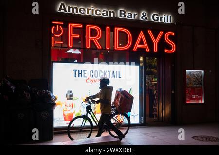 Madrid, Espagne. 30 mars 2024. Un livreur à la demande transportant un vélo passe devant la chaîne de restaurants décontractés américaine TGI Fridays. (Crédit image : © Xavi Lopez/SOPA images via ZUMA Press Wire) USAGE ÉDITORIAL SEULEMENT! Non destiné à UN USAGE commercial ! Banque D'Images