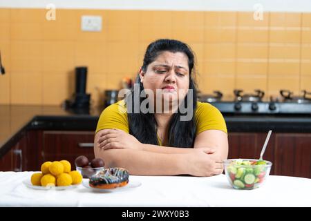 Triste surpoids graisse indienne femme confus qui de choisir entre salade saine ou dessert, laddu placé sur un côté d'une table dans la cuisine. Régime St Banque D'Images