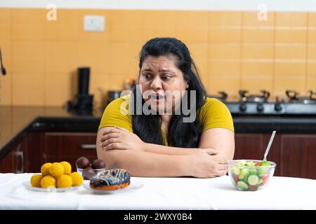 Grosse femme indienne en surpoids excitée de voir des beignets et laddu ou dessert. Salade de légumes placé sur un côté d'une table dans la cuisine.Dieting lutte conc Banque D'Images