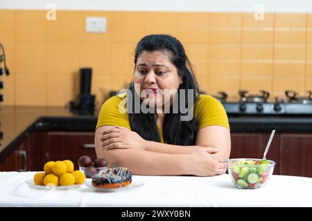 Grosse femme indienne en surpoids excitée de voir des beignets et laddu ou dessert. Salade de légumes placé sur un côté d'une table dans la cuisine.Dieting lutte conc Banque D'Images