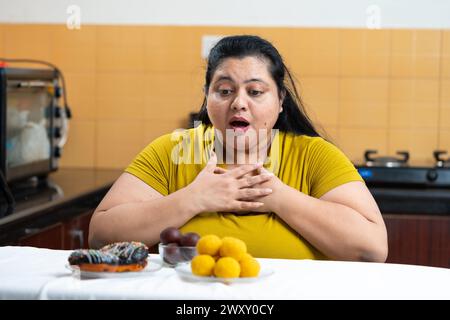 Grosse femme indienne en surpoids s'excitant de voir des beignets et laddu ou dessert sur une table dans la cuisine. Manger malsain, concept d'envie. Banque D'Images