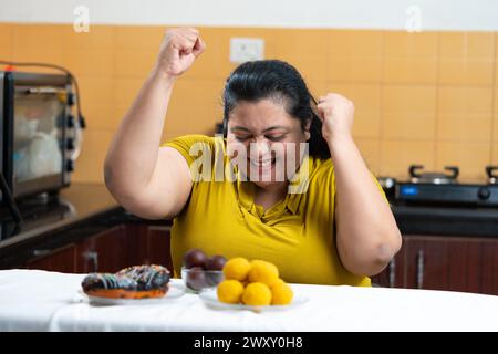 Grosse femme indienne en surpoids s'excitant de voir des beignets et laddu ou dessert sur une table dans la cuisine. Manger malsain, concept d'envie. Banque D'Images