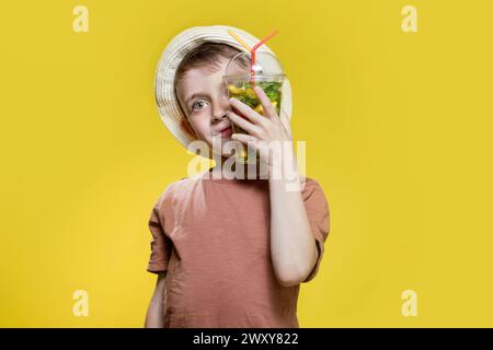 Garçon mignon au panama buvant un cocktail mojito de tasse en plastique sur fond de studio jaune. Banque D'Images