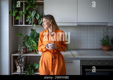 Femme souriante insouciante buvant du thé dans la cuisine parmi les plantes d'intérieur, regardant par la fenêtre. Vie lente. Banque D'Images