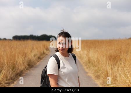 Jeune femme avec sac à dos debout sur la route dans le champ, regardant la caméra Banque D'Images