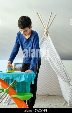 Un jeune garçon se concentre sur le repassage d'un morceau de tissu sur une planche à repasser jouet, avec un tipi en arrière-plan, illustrant l'imagination de l'enfance et le pla Banque D'Images