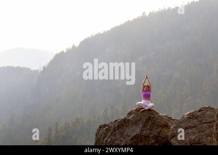 Un personnage est silhouetté contre un paysage de montagne brumeux, assis dans une pose de yoga sur une falaise avec les mains en position de prière Banque D'Images