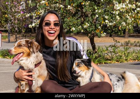 Une femme joyeuse en lunettes de soleil est assise avec deux chiens bergers australiens entourés de fleurs en fleurs dans un parc ensoleillé Banque D'Images