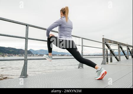 Une femme active étire sa jambe contre une balustrade sur une promenade en bord de mer avec un ciel nuageux et des montagnes en arrière-plan Banque D'Images