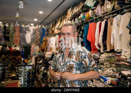 Un commerçant joyeux se tient dans un magasin de vêtements dynamique, entouré d'une gamme de vêtements et d'accessoires à motifs Banque D'Images