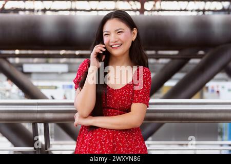 Une jeune femme joyeuse dans une robe rouge s'engage dans une conversation sur son téléphone portable, appuyée sur une balustrade métallique avec une infrastructure urbaine floue b Banque D'Images